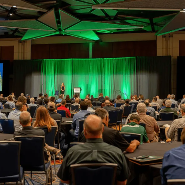A large audience sits at tables in a conference room, attentively listening to a speaker on stage with green lighting.