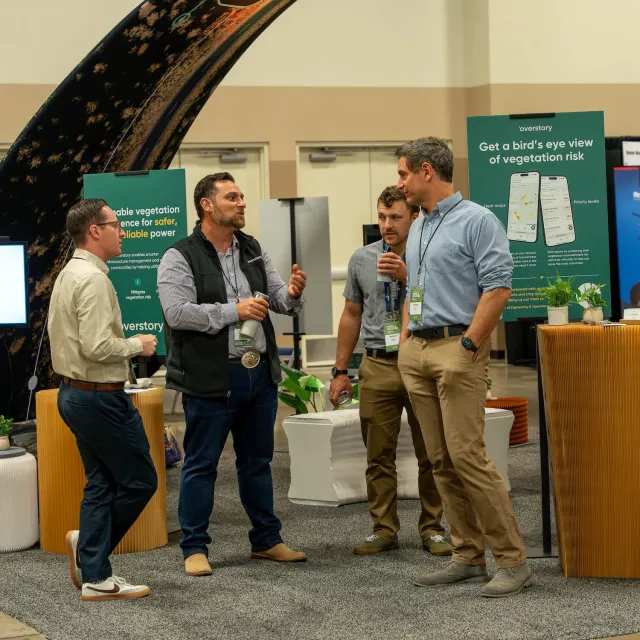 A group of four men engage in conversation at a trade show booth, surrounded by plants and promotional materials about vegetation risk.
