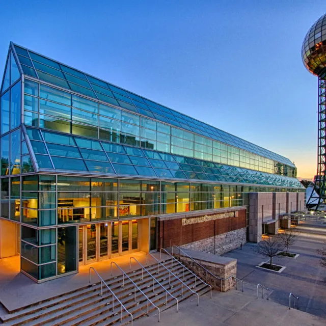 A modern glass building, the Knoxville Convention Center, stands alongside a towering sphere structure, illuminated at dusk.