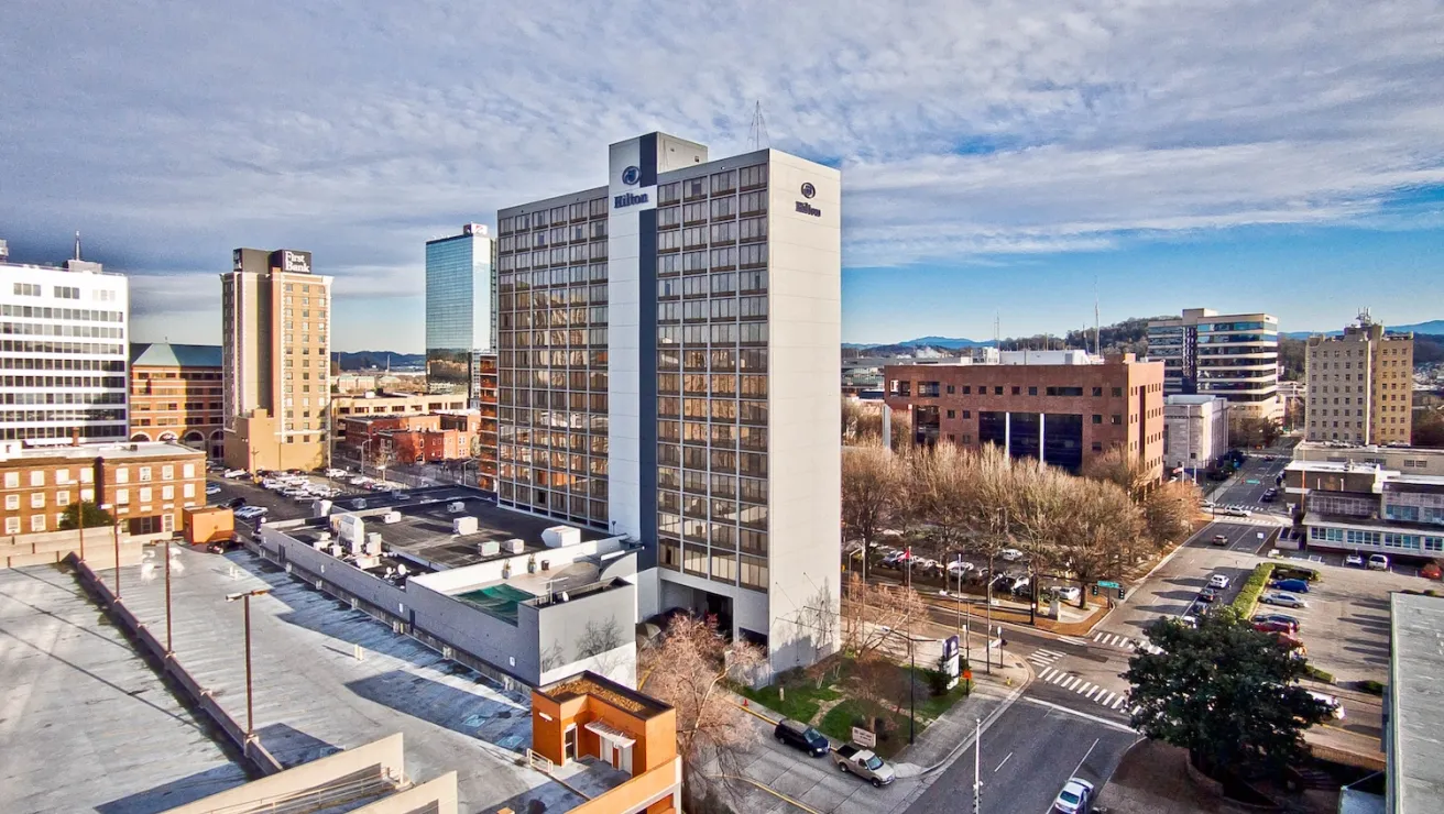 Aerial view of downtown Knoxville, featuring a Hilton hotel and various buildings under a clear blue sky with distant mountains.