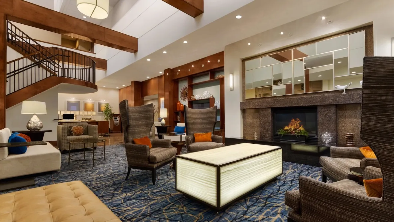 Modern hotel lobby featuring cozy seating, elegant lighting, a fireplace, and a stylish staircase, all on a patterned blue carpet.