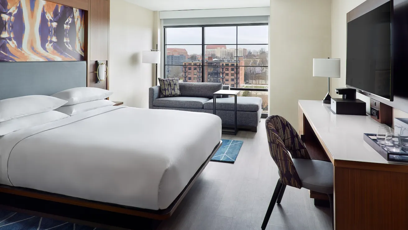 Modern hotel room featuring a king bed, seating area, desk, and large window with city views. Elegant decor and cozy atmosphere.