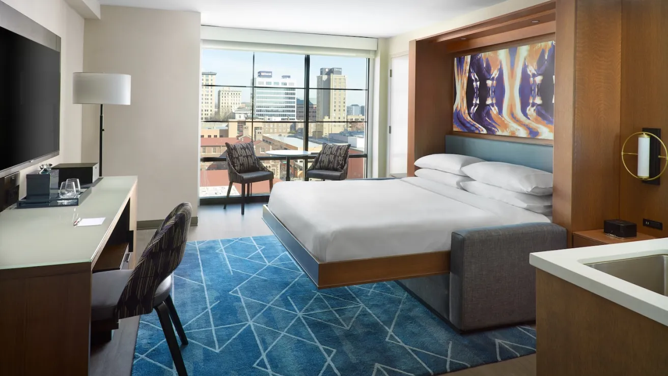 Modern hotel room featuring a queen bed, sleek desk, cozy seating, vibrant artwork, and large windows with city views.