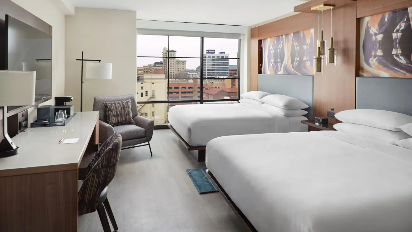 A modern hotel room featuring two queen beds, a desk, and city views through large windows. Stylish decor and comfortable seating included.