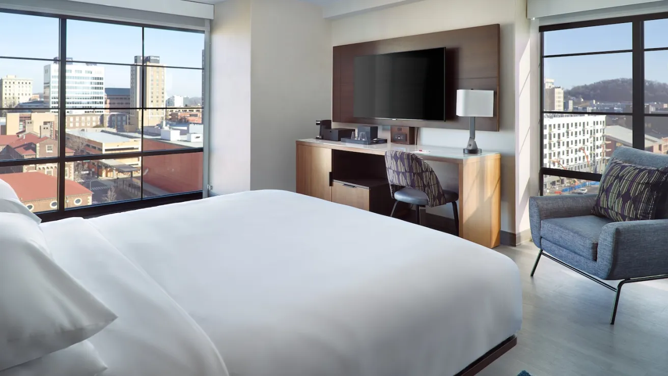 Modern hotel room featuring a king bed, desk area, lounge chair, large window with city views, and a wall-mounted TV.