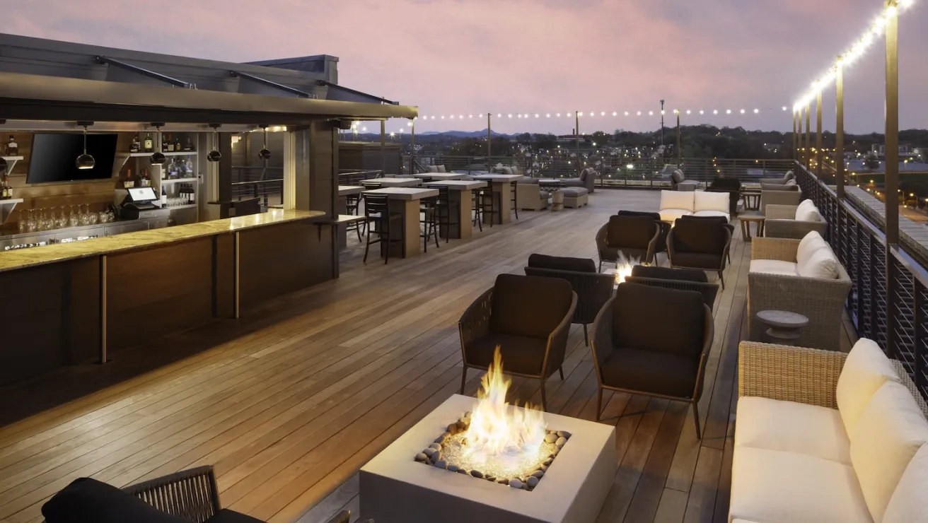 Modern rooftop lounge with a fire pit, sleek bar area, and cozy seating, set against a sunset sky and cityscape.
